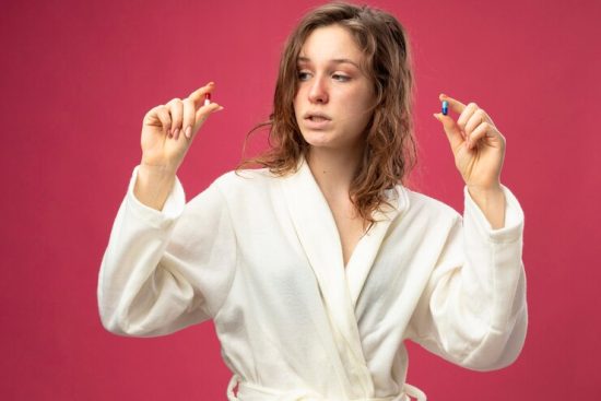 pensive-young-ill-girl-wearing-white-robe-holding-looking-pills-isolated-pink_141793-86350