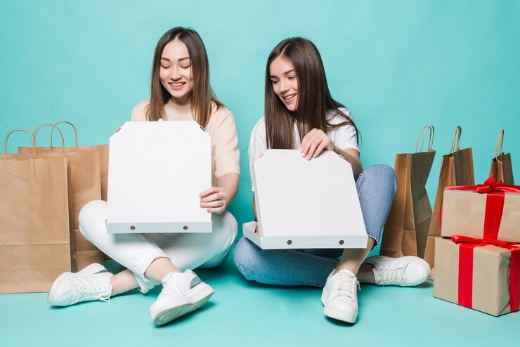 smiling-young-two-girls-sitting-floor-shopping-bags-gift-open-pizza-turquoise-wall_231208-11845